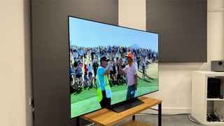65-inch LG C4 TV photographed at a slight angle on a wooden stand. On the screen is an image of two golfers congratulating one another.