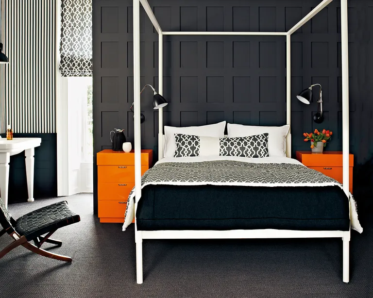 A bedroom with black panelled walls, white four poster bed, black and white soft furnishings and orange side tables