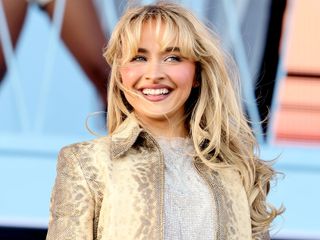 Sabrina Carpenter smiles and looks away from the camera wearing a beige jacket