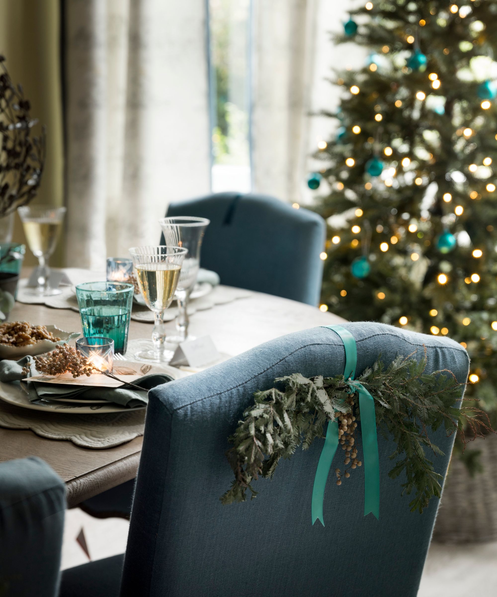 Christmas table with foliage on the chair back and green and clear glassware with Christmas tree in the background