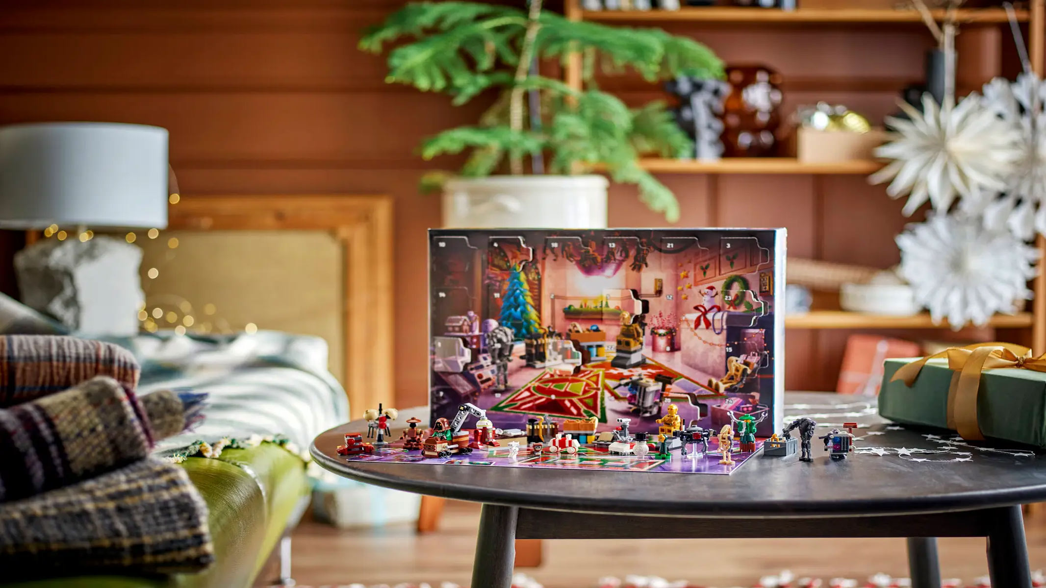 An advent calendar with brick-built droids laid out is infront of a festive backdrop, on a table in a living room with a green plant in the background.