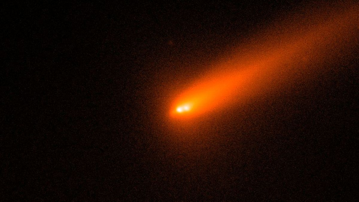 Se muestran dos puntos brillantes de luz blanca sobre un fondo oscuro que representa el núcleo de un cometa fracturado, rodeados por una coma roja brumosa y una cola que se aleja hacia la parte superior derecha de la imagen.