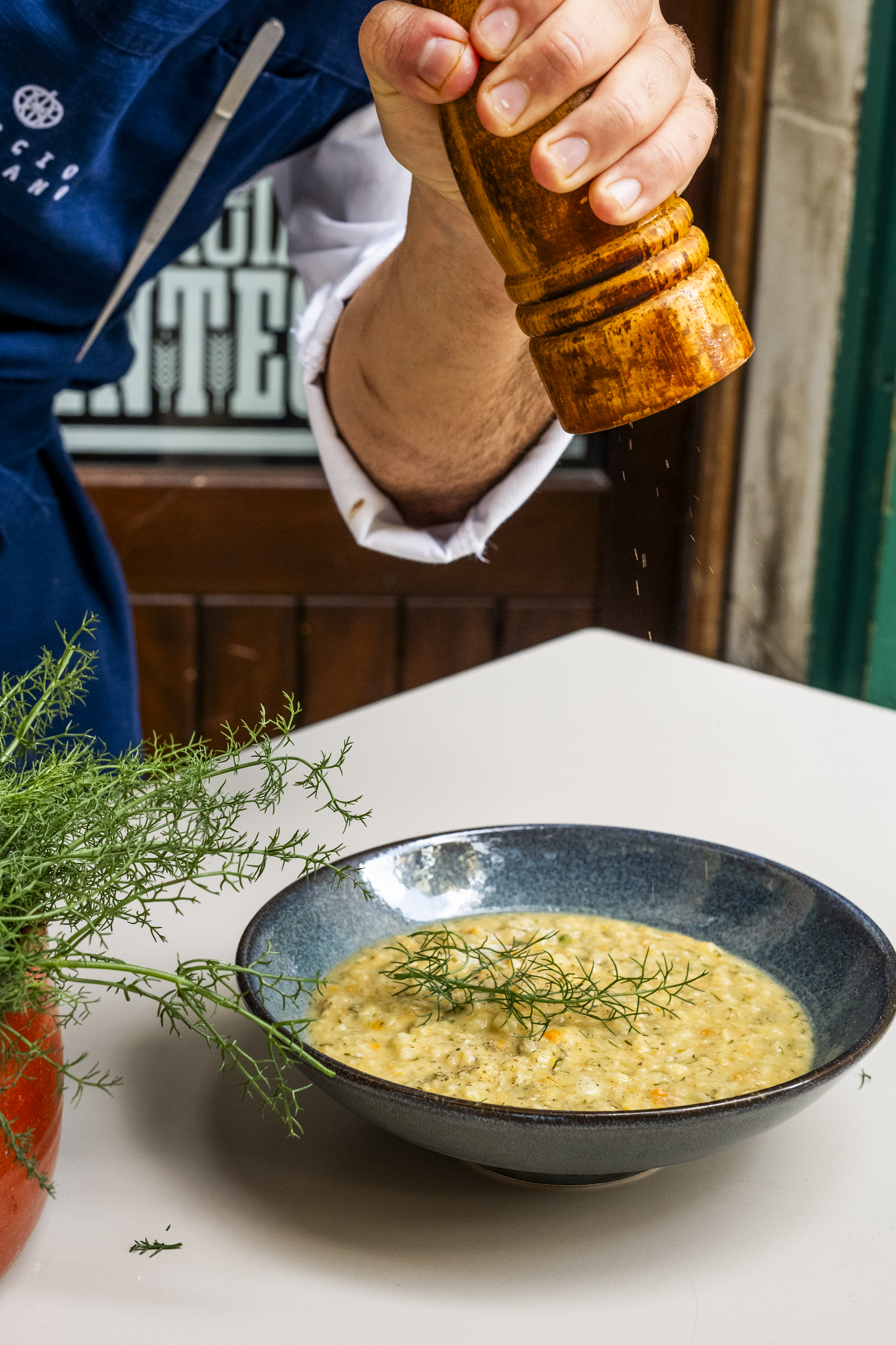 The hand of a chef is captured grinding some pepper onto a risotto dish served in a glazed ceramic bowl in blue.