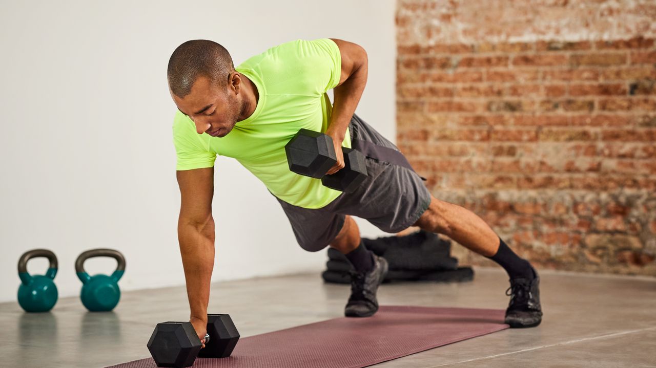 A man performs a renegade row in a sparse room with dumbbells on a yoga mat. He is in a push-up position, with dumbbells in each hand. One is on the floor, the other is close to his torso as he has pulled back his elbow to lift it. 