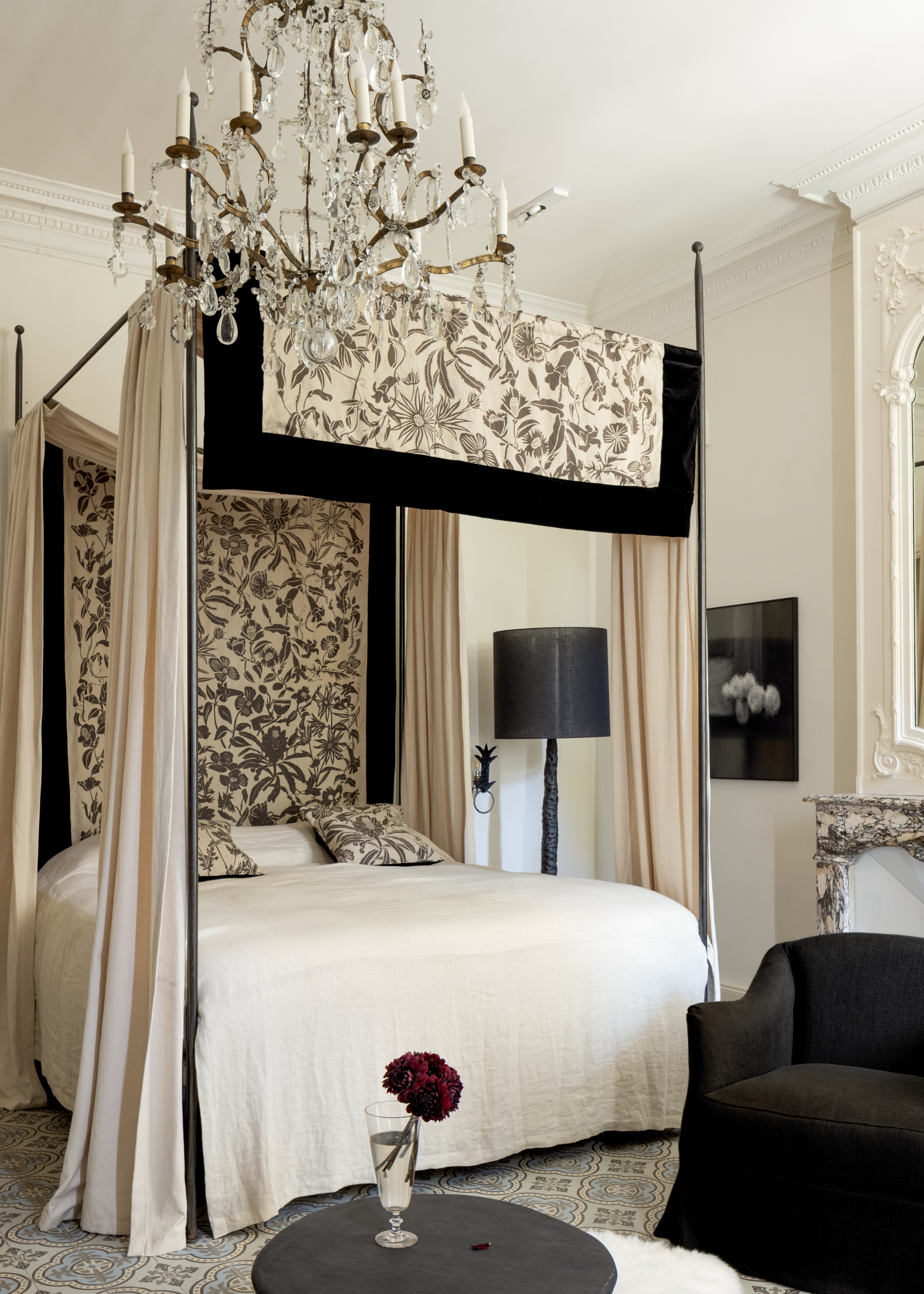 A white bedroom with a patterned canopy, a white bedscape, a tall floor lamp, a black armchair, and a side table with a vase
