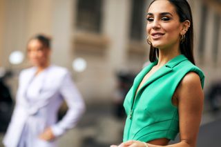 a guest at milan fashion week fall 2023 wears a green sleeveless blazer and gold hoop earrings and ear cuffs smiles