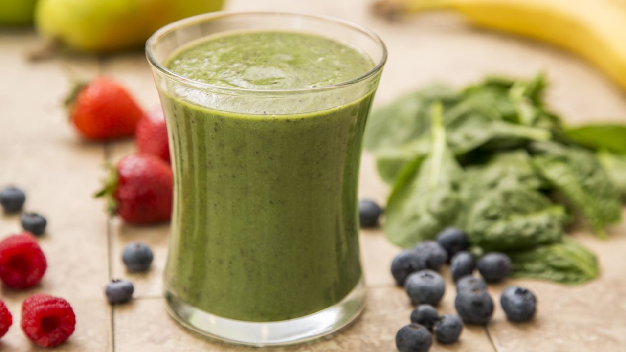 A green smoothie in a glass surrounded by spinach, blueberries, and strawberries on a table