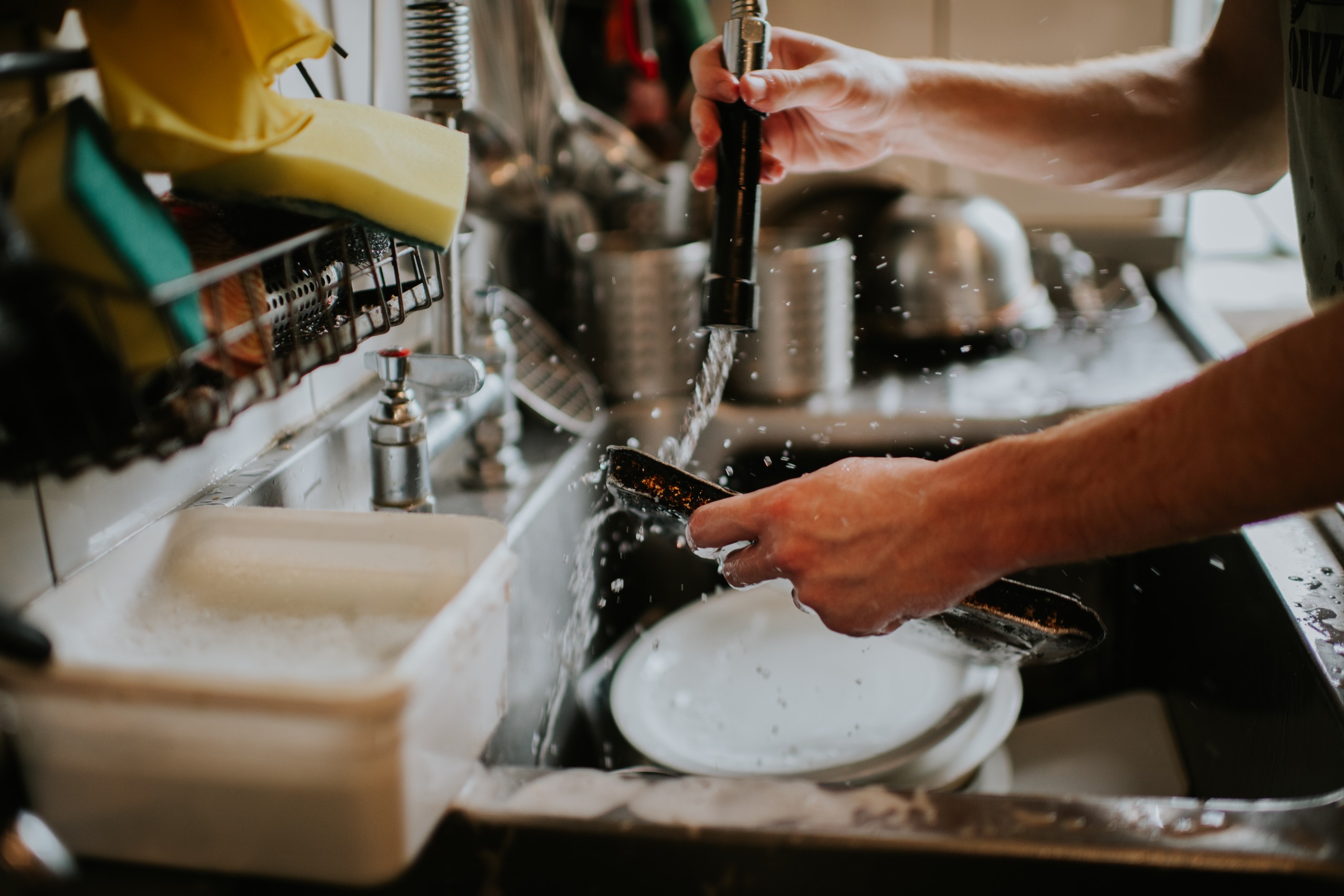A man washes a baking sheet in his sink, which is filled with other dirty dishes.