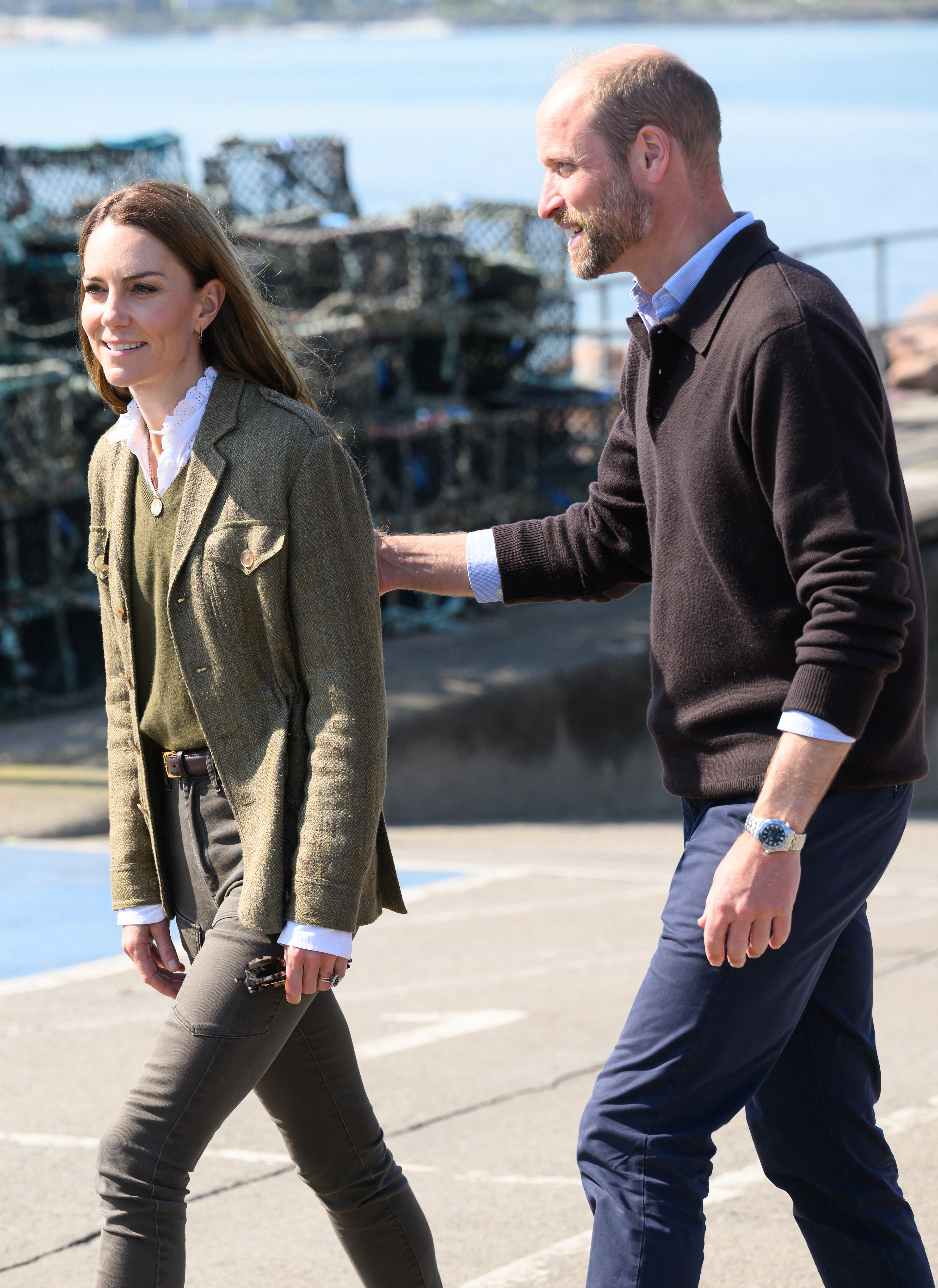 Kate Middleton wearing a green blazer walking next to Prince William, in a a brown sweater, putting his hand on her back