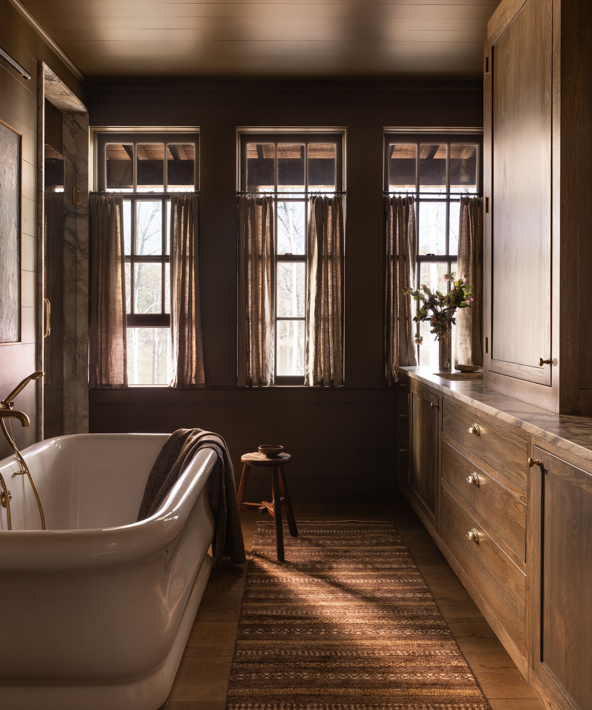 Moody, rustic bathroom with dark walls, three tall windows with linen cafe curtains, a white clawfoot tub, and extensive warm wood cabinetry with marble counters.
