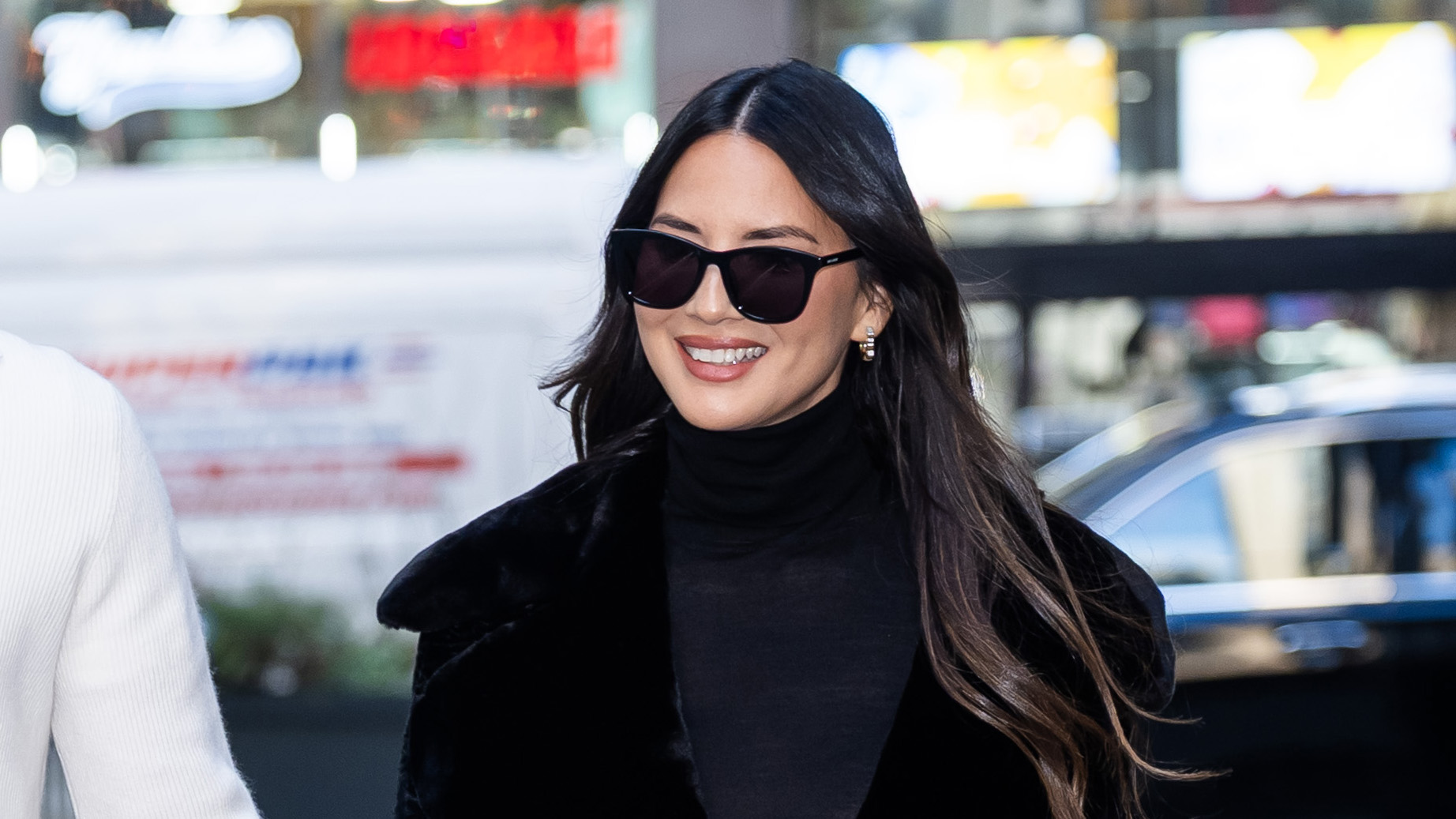 Olivia Munn with John Mulaney wearing a black coat and turtleneck, stovepipe blue jeans, and black pumps.