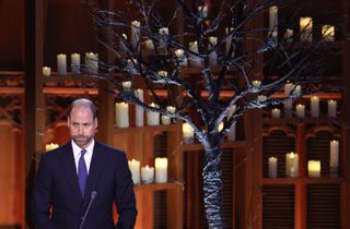 Prince William wearing a dark suit and tie standing in front of a tree and lit candles in a dark room on stage