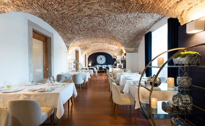 Inside look of the Ânfora restaurant. Rows of tables covered with white linen surrounded by beige chairs. White walls and lowered exposed brick ceilings.