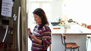 Woman looking at phone next to fridge in kitchen, wondering why she's dieting but not losing weight
