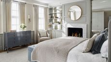 A neutral bedroom with large double bed, cream rug, blue chest of drawers, glass-backed shelves, a round mirror, white closet, and a white marble fireplace with a fire burning.