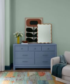 corner of bedroom with pastel green walls, light blue dresser with mirror, artwork and vase of flowers on top