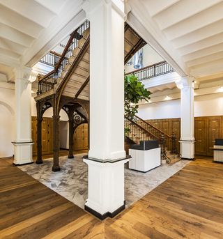 Reception desk at the bottom of a grand staircase. White pillars feature, as well as wooden floors and wall panels