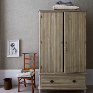 A wooden wardrobe with a chair next to it set against a textured fabric wallpaper in grey