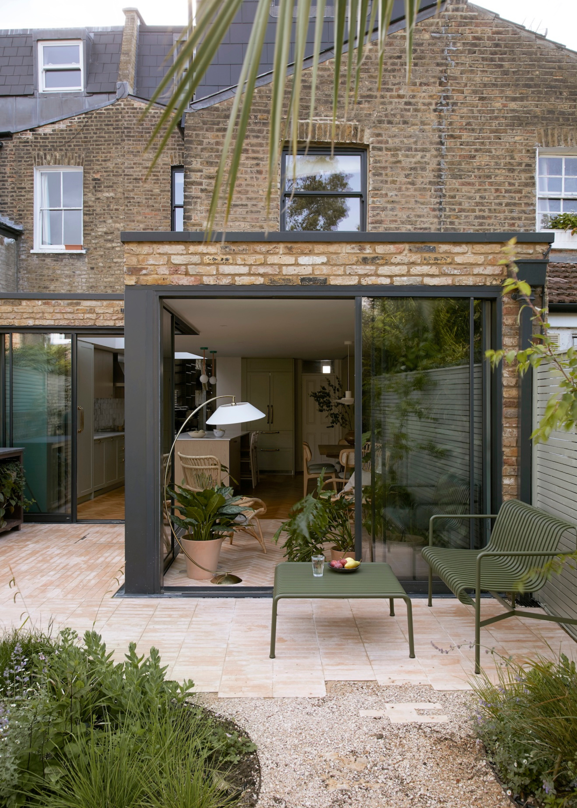 An outdoor living room with a green bench and table leading to an indoor plant room