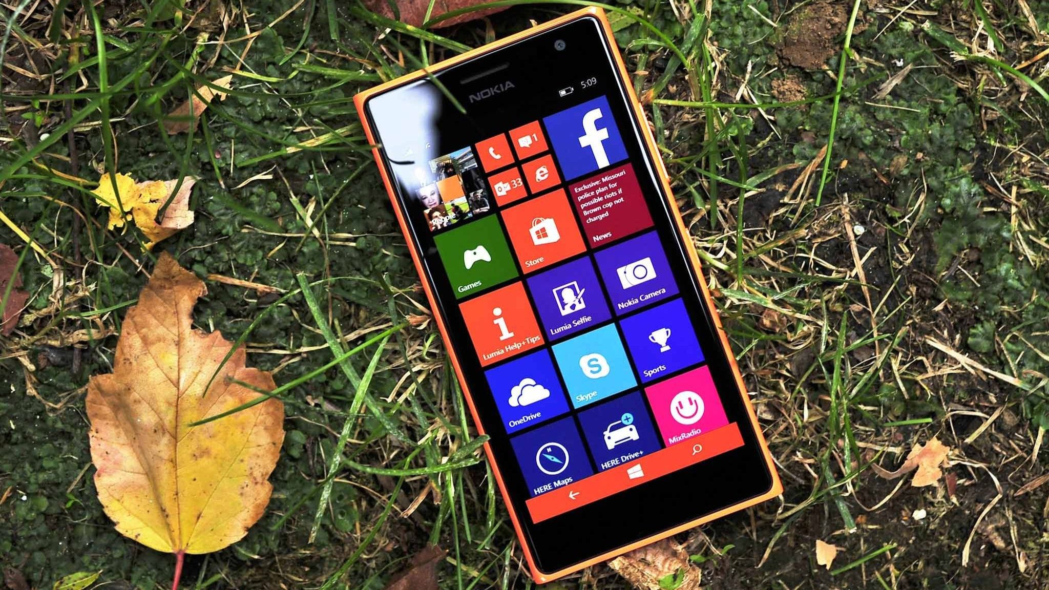 A smartphone with a colorful app tile interface lies on grass surrounded by autumn leaves. The bright screen contrasts with the earthy background.