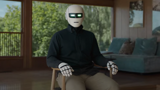 A humanoid robot wearing an oxford shirt sitting in a chair in a house. Its greenish rectangular eyes look directly at the camera.