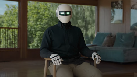 A humanoid robot wearing an oxford shirt sitting in a chair in a house. Its greenish rectangular eyes look directly at the camera.