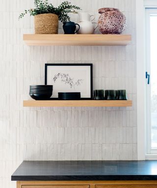 white kitchen with black countertops, two floating shelves with plates, plant, vases, white pearlescent wall tiles