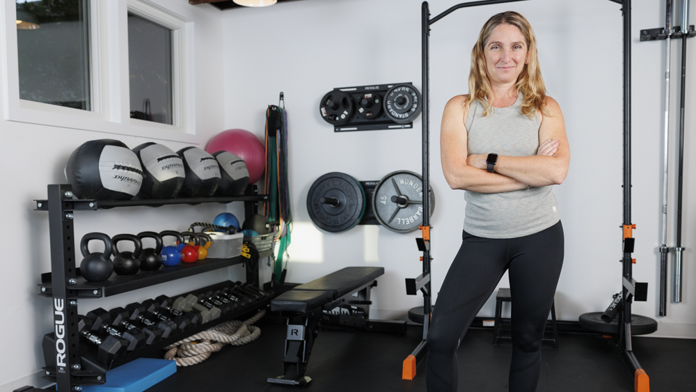 Woman in gray tank top and black leggings stands with her arms folded, a range of exercise equipment behind her