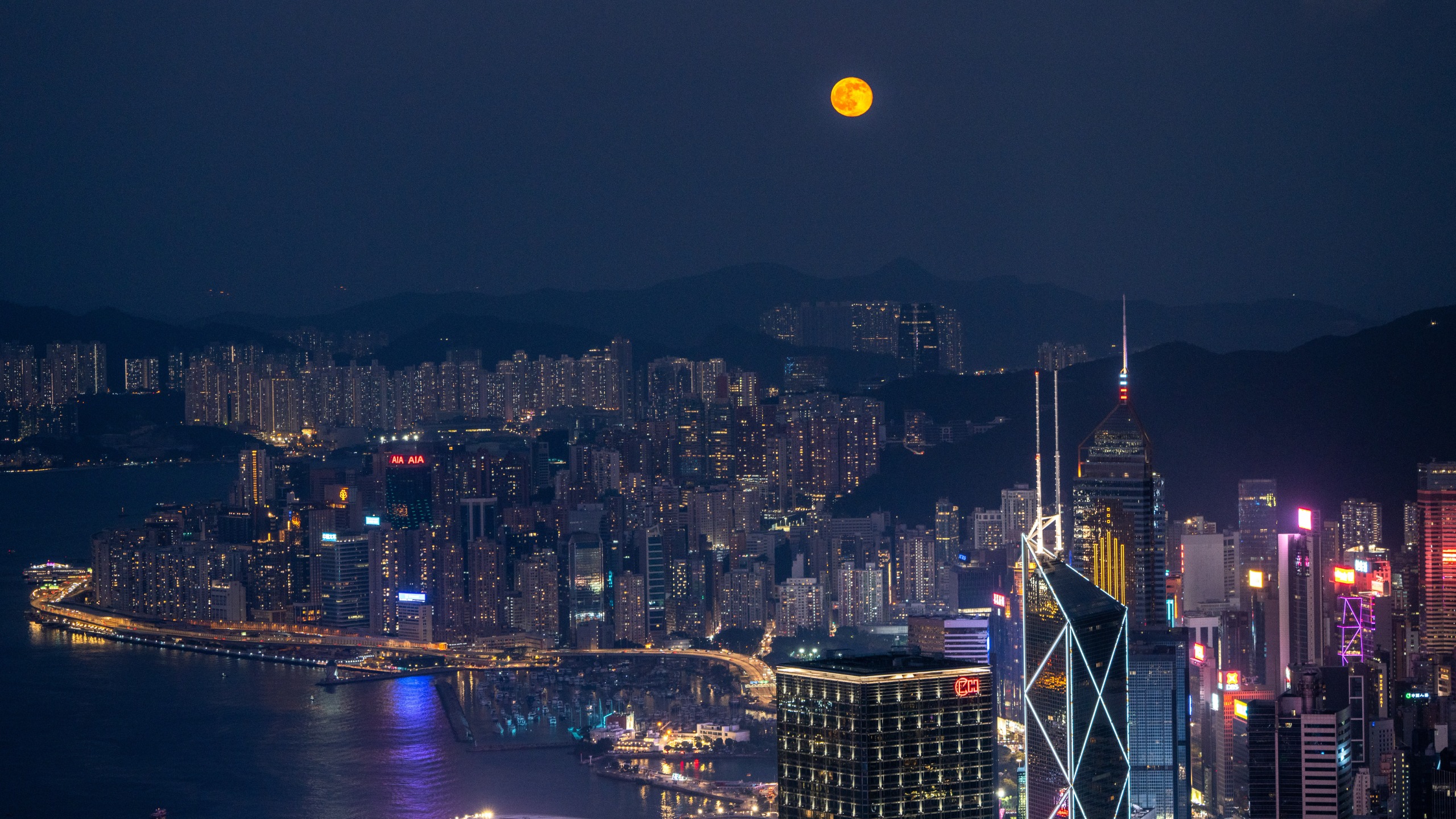 The bright full moon hangs above the illuminated skyscrapers surrounding Victoria Harbour in Hong Kong at night.