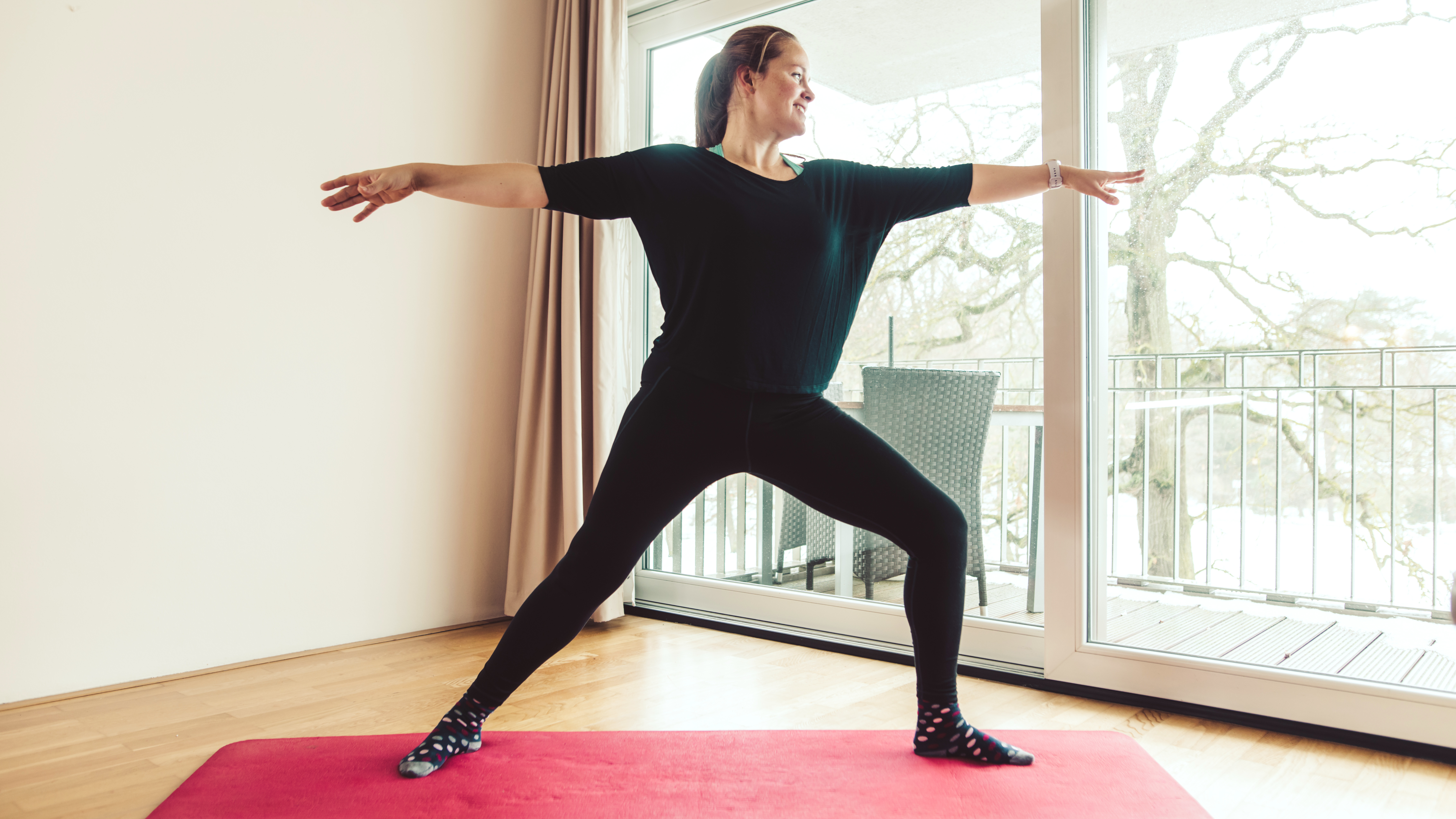 A woman practice a warrior two yoga pose on a mat; she is in a lunge with her left foot forward and right foot back. Her arms are extended at shoulder height.