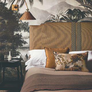 A bedroom with a mural wallpaper and a bed with a brown fabric headboard matching the rattan wall light and cushions and bedspreads on the bed