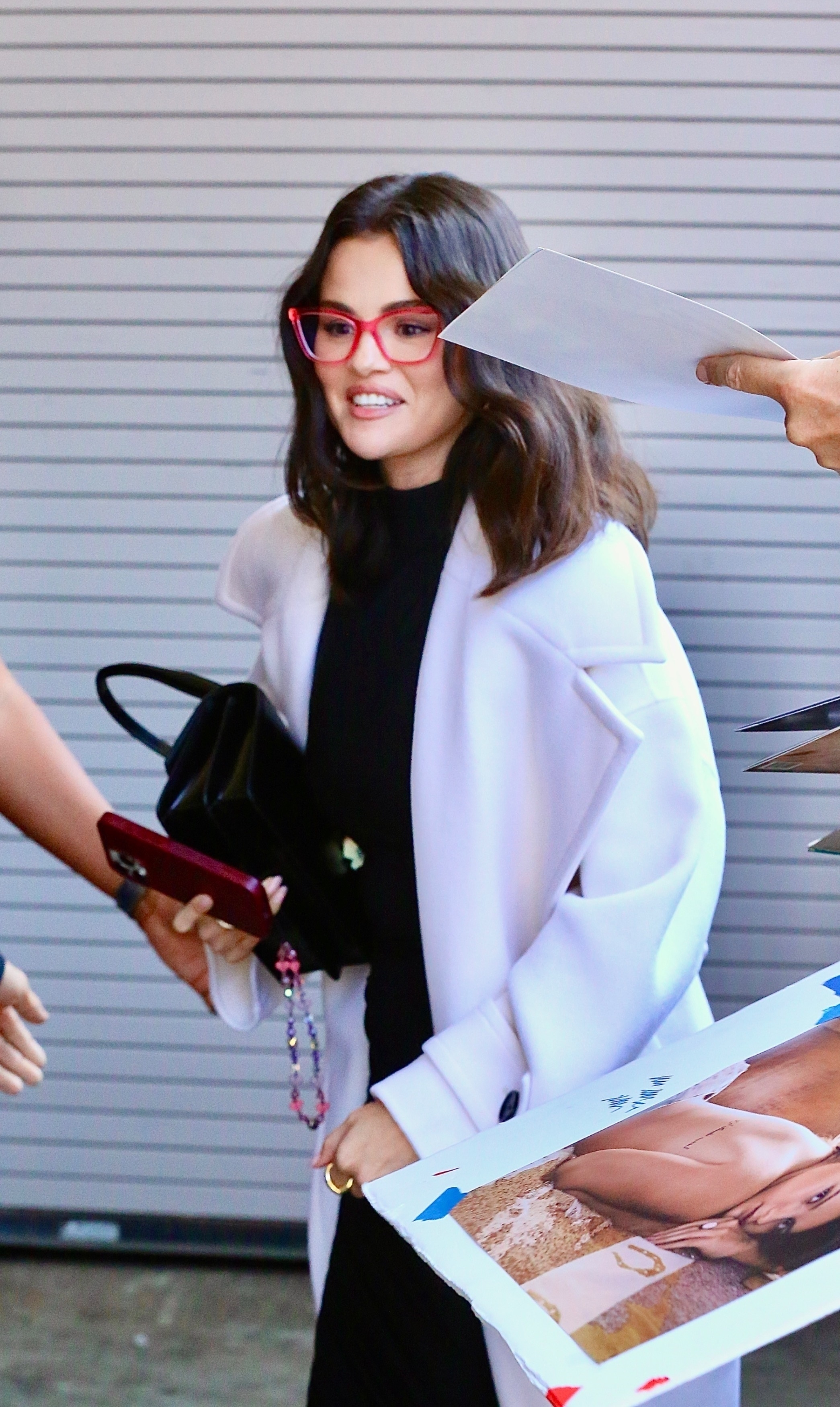Selena Gomez wearing heeled flip-flops, a black dress, a white coat, and red glasses