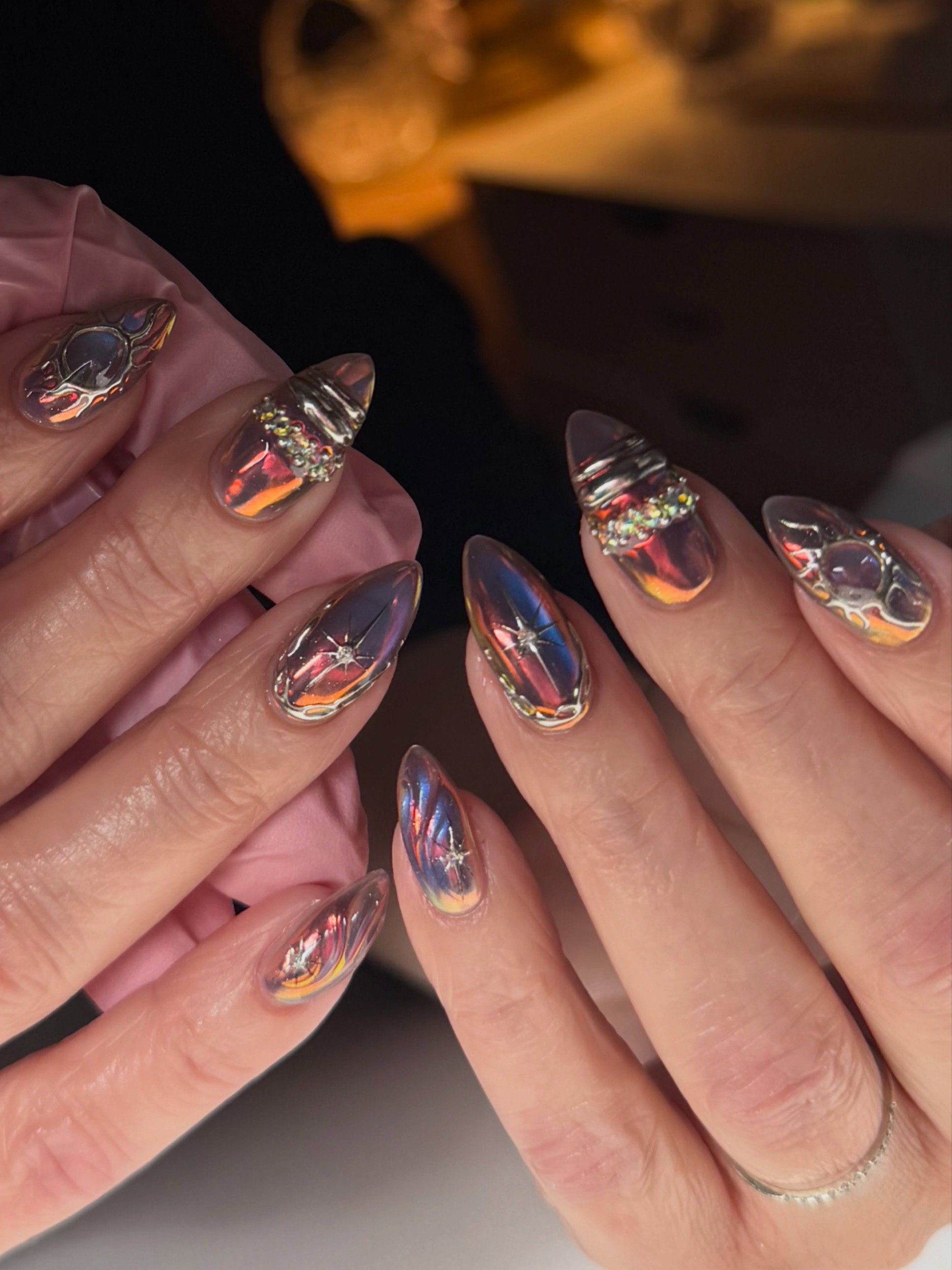 Close-up of long, pointed nails with colorful chrome designs, metallic details, and rhinestones