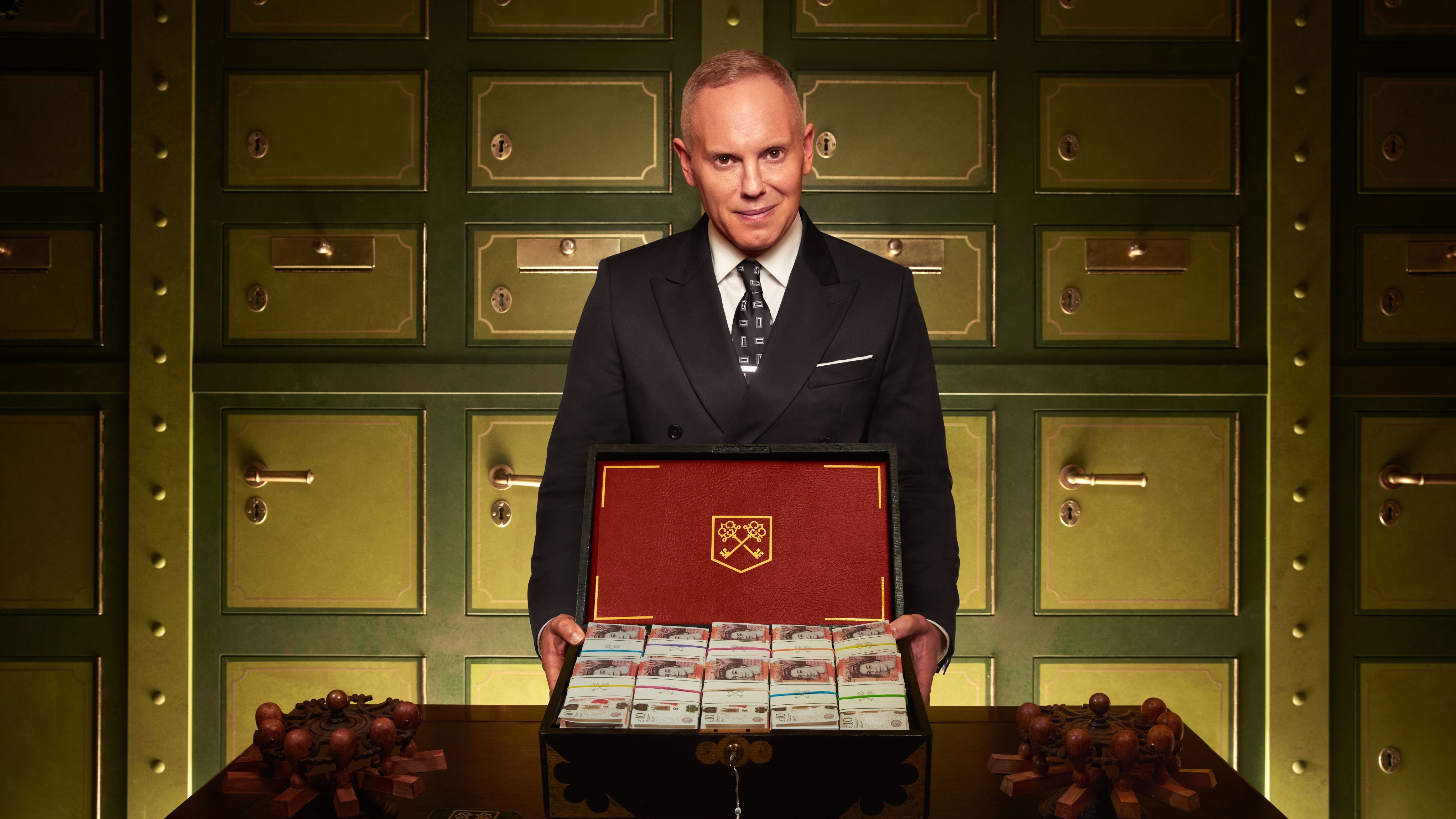 Robert Rinder as The Executor holding a suitcase full of cash open in front of him.