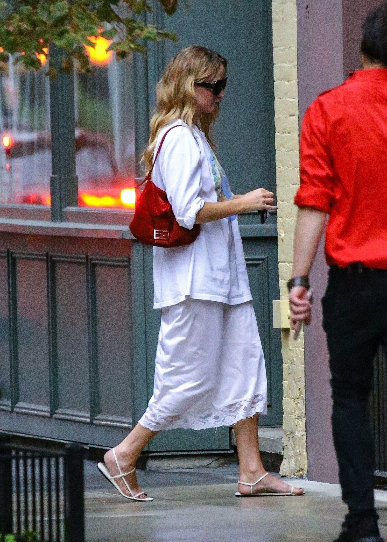 Jennifer Lawrence enters into a building wearing a white lace-trim slip skirt with a white baggy t-shirt and white strappy sandals. She wears a red suede bag over her shoulder.