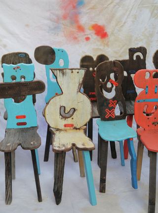 View of multiple Folk chairs by Serban Ionescu - red, blue, cream and dark wood chairs with backrests that feature carved whimsical expressions pictured in front of a paint stained piece of fabric