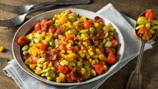 Bowl of homemade succotash sitting on a wooden table on top of a napkin with two forks and a spoon beside it 