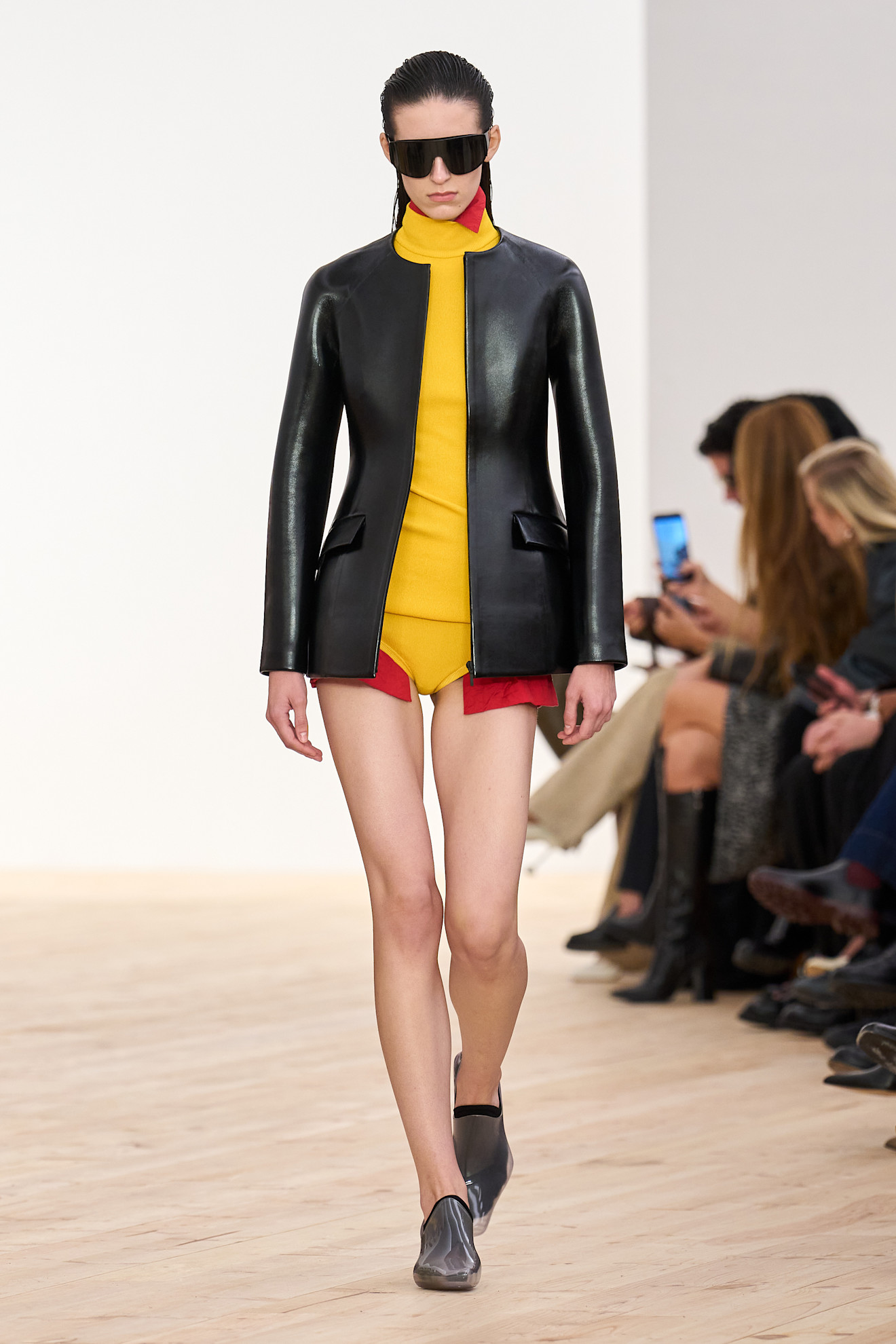Layering staples for spring 2026 is shown in a photo from Loewe's runway show, with a model walking while wearing a bright red oversized button-down shirt layered under a yellow turtleneck sweater and yellow briefs, with a black leather jacket layered on top. The rest of the look is styled with black aviators and clear pointed loafers styled with black socks