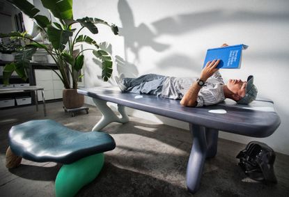A portrait of Daniel Arsham lying down on a table, reading a blue book
