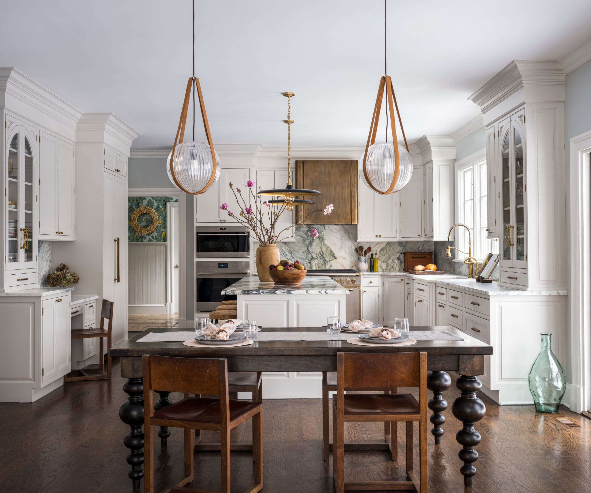 A large kitchen with white shaker cabinets, marble countertops, brass hardware, and a dark wooden dining table