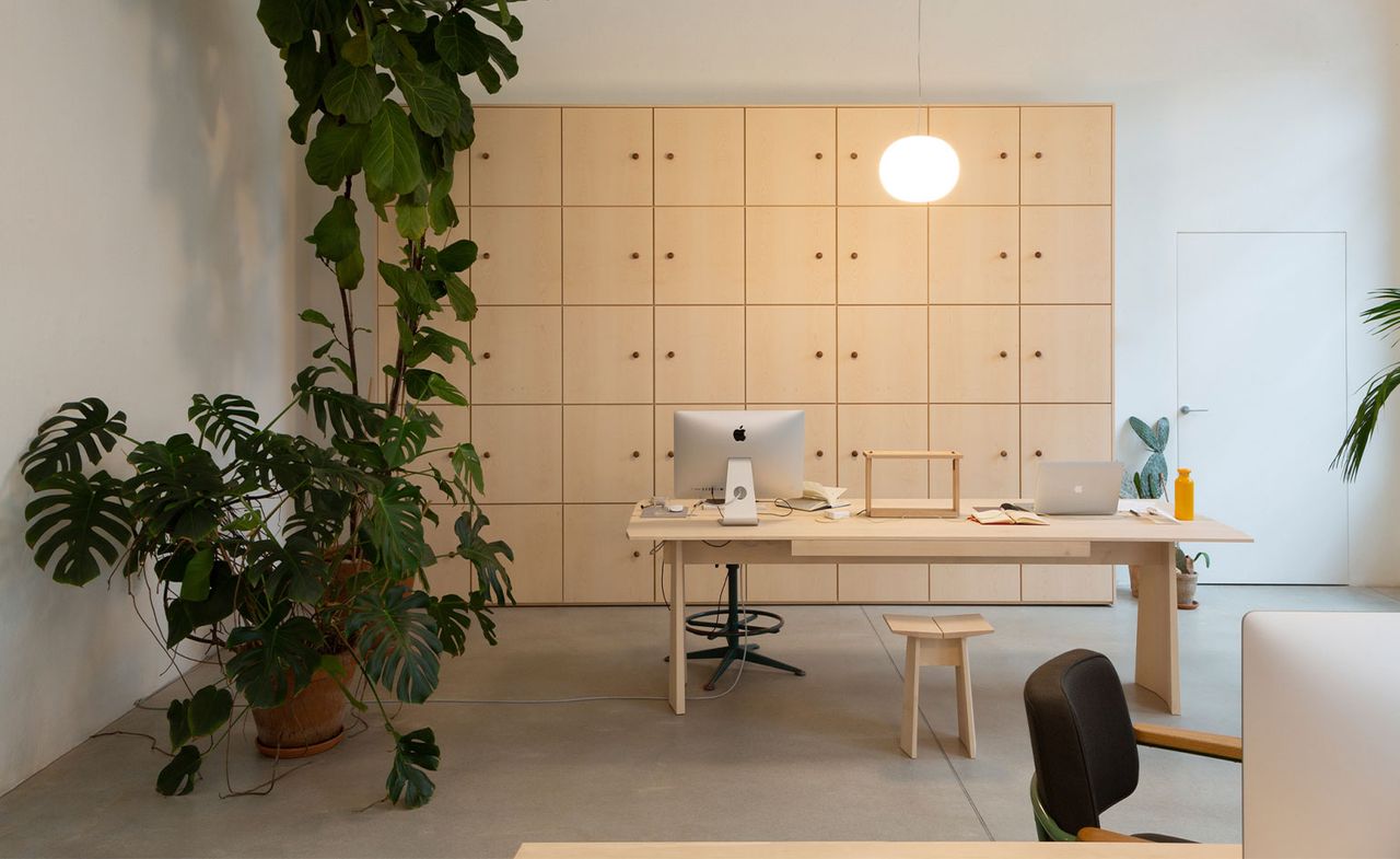 Interior view of the Formafantasma Milan studio featuring grey floors, white walls, a pendant light, custom built light wood office furniture and large green plants in pots