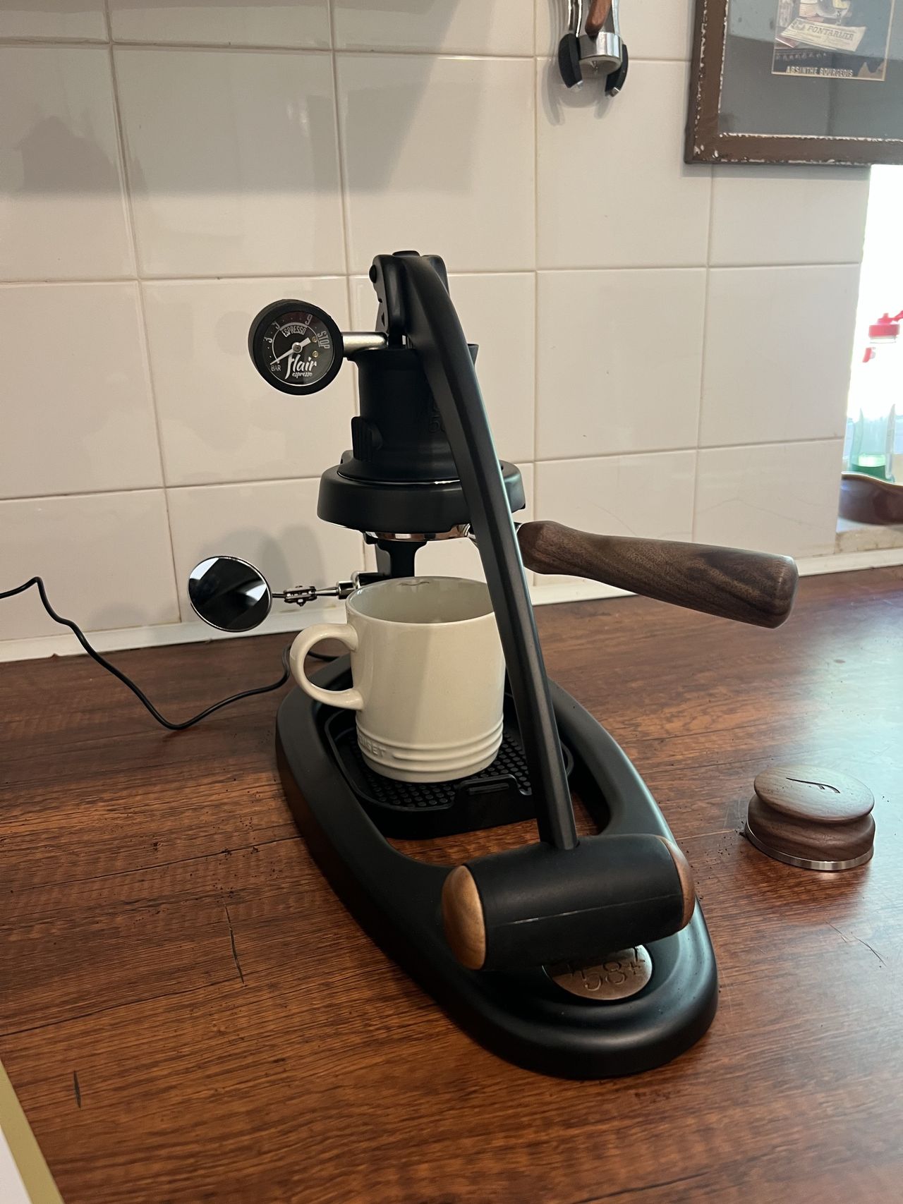 A white coffee mug under the brew head of a black and wood manual coffee machine sitting on a wooden countertop in a kitchen with white tiles on the wall.