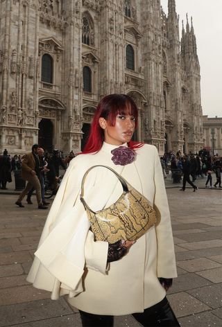 A snakeskin purse is shown in a street-style photo of a woman standing outside wearing a white cape dress with black thigh-high patent leather boots and a yellow snakeskin shoulder bag