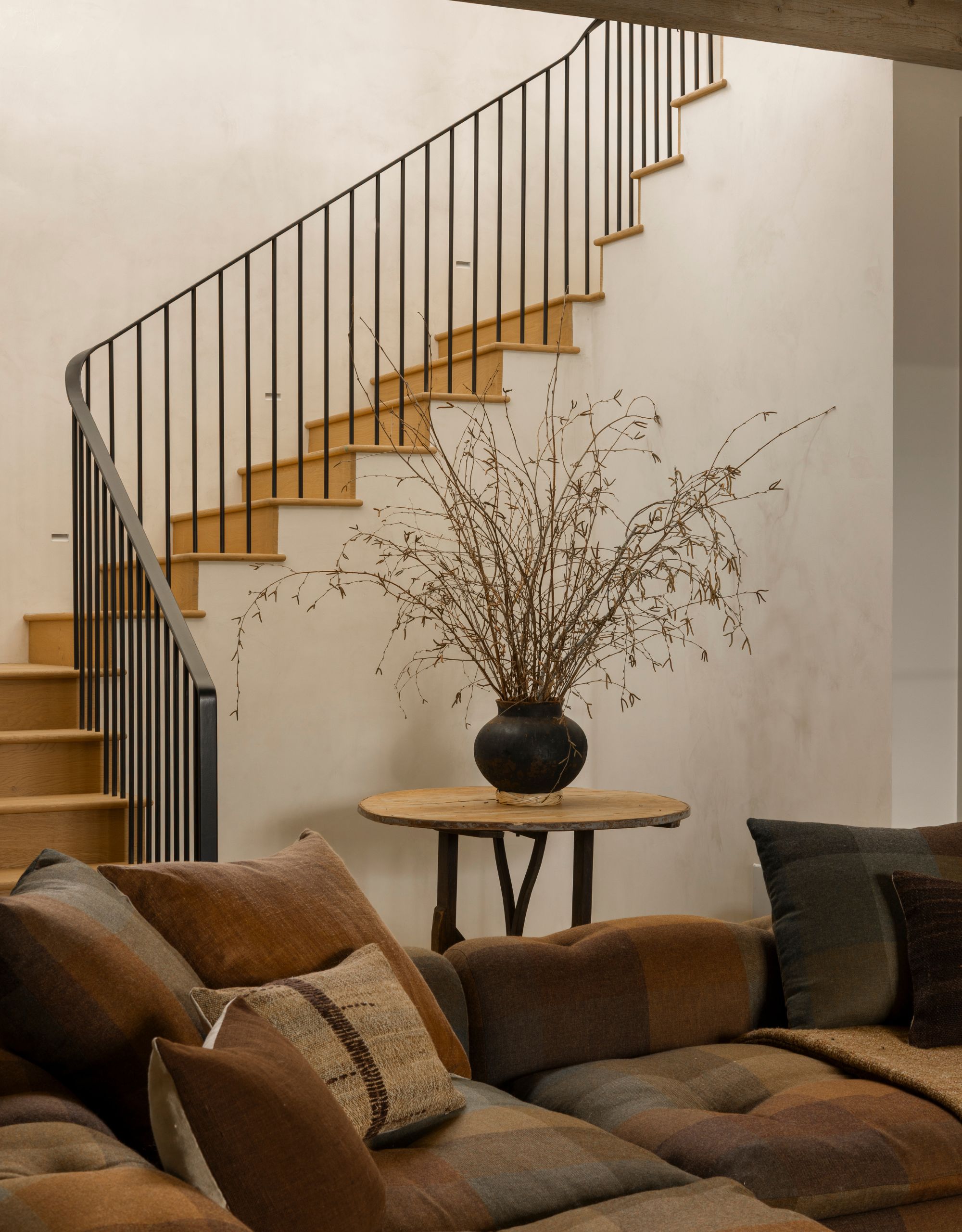 A staircase leads down to a living room with a plaid style sofa with lots of cushions and a round table sits at the bottom of the stairs with a large vase of foliage on top