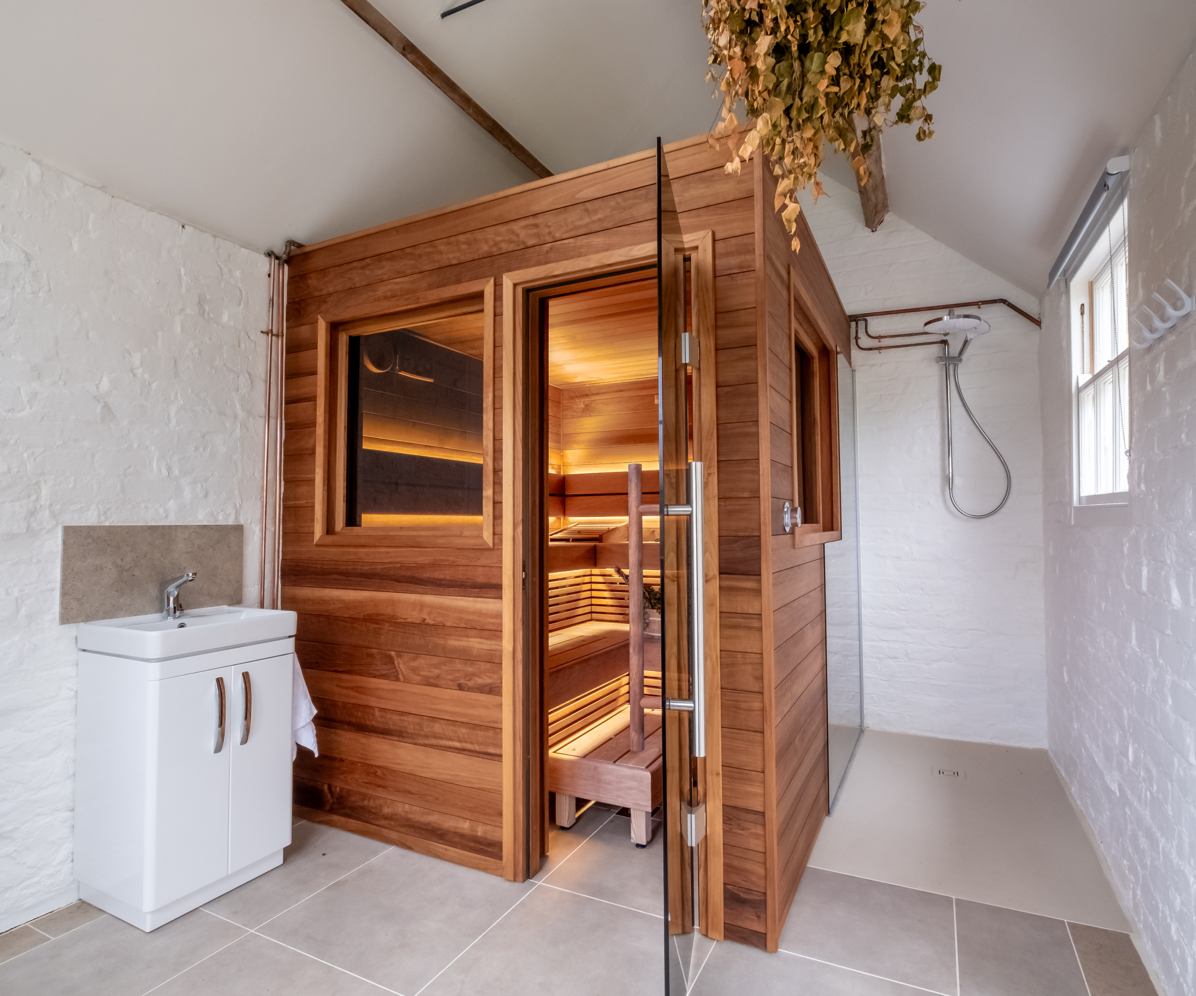 wooden sauna inside building with white painted brick walls and tiled floor, small sink and vanity unit to one side of sauna
