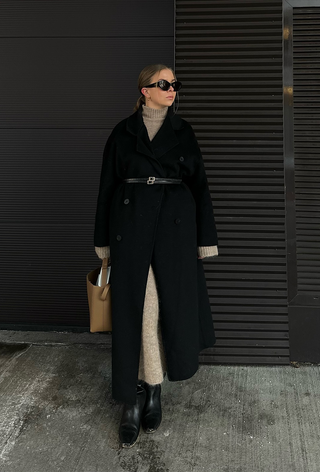 what to wear in 30-degree temperatures shown in a photo of a woman wearing a black coat over a tan maxi sweater dress styled with a black belt, black knee-high boots, and a tan tote bag