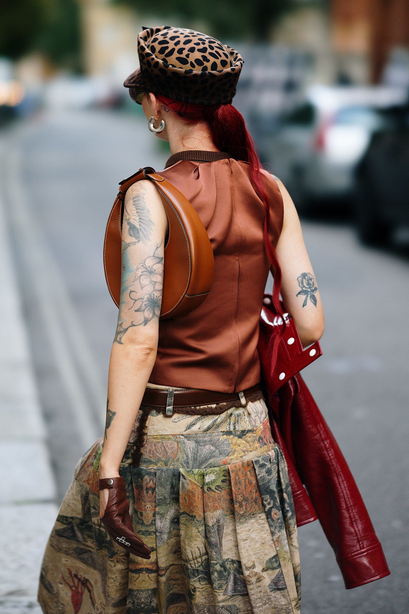 a tattooed woman in a red vest and patterned green skirt with red hair in a ponytail facing away from the camera