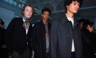 Three male models wearing looks from Margaret Howell's collection. One model is wearing a white top, black cardigan, grey trousers and dual coloured scarf tied around his neck. Another model is wearing a grey top, black coat and dual coloured scarf. And the third model is wearing a white top, black trousers and a blue coat