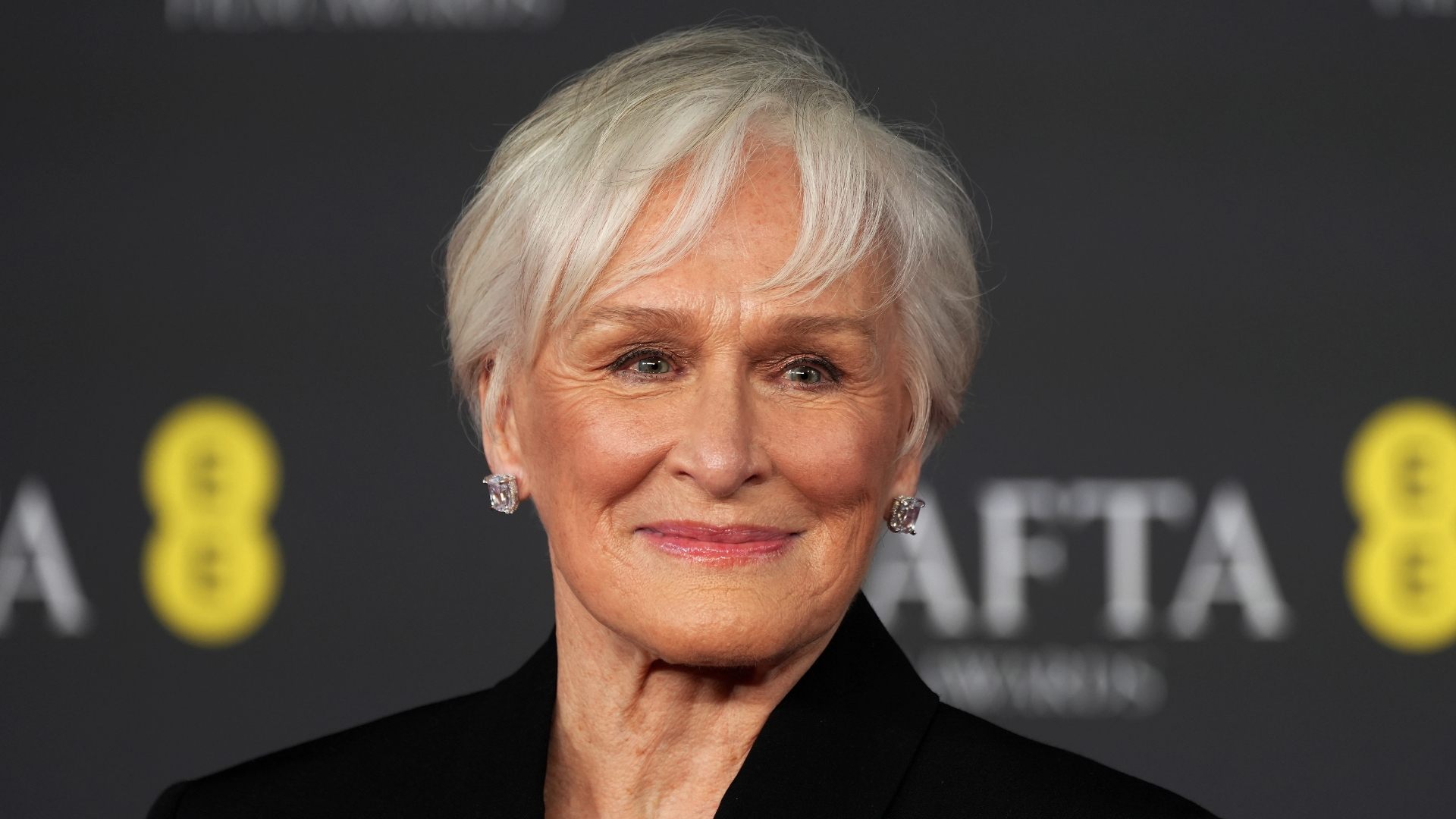 Image of Glenn Close smiling with grey hair pixie cut, rosy pink lipstick and square diamond earrings