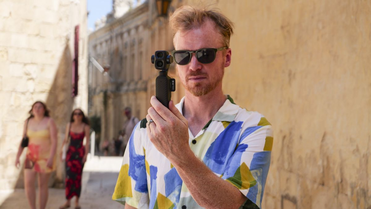 Man in bright summer short and shades, in an old town on a sunny day, holding the DJI Osmo Pocket 4P vlogging camera
